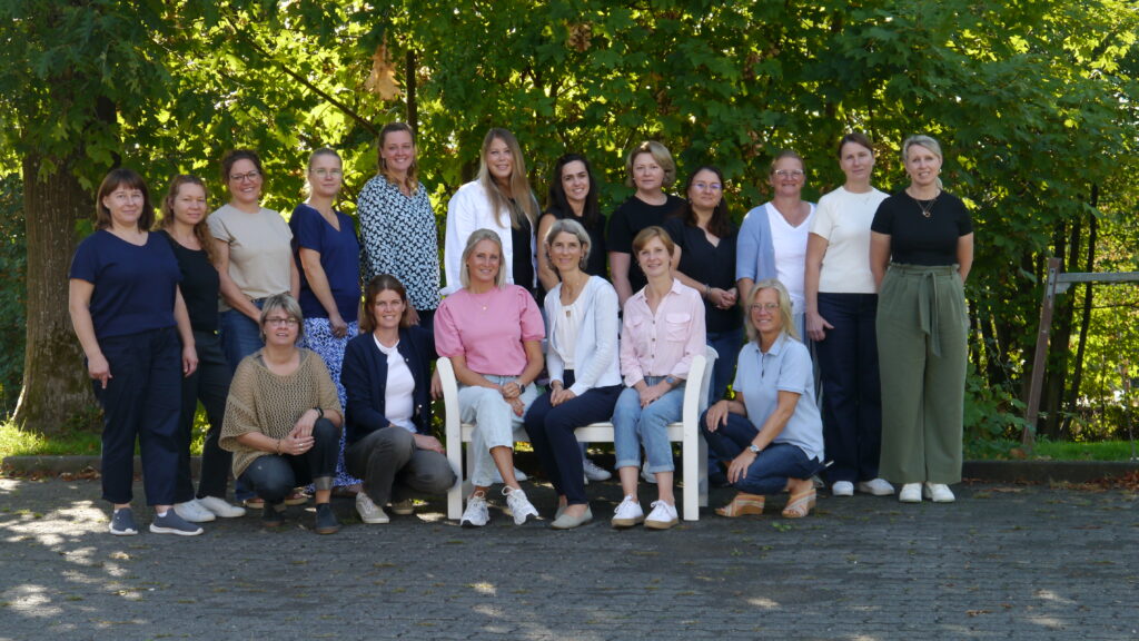 Eine Gruppe von 19 Erwachsenen, überwiegend Frauen, posiert im Freien vor grünen Bäumen für ein Foto. Einige sitzen auf Stühlen, während andere hinter ihnen stehen. Alle sind leger gekleidet und lächeln.